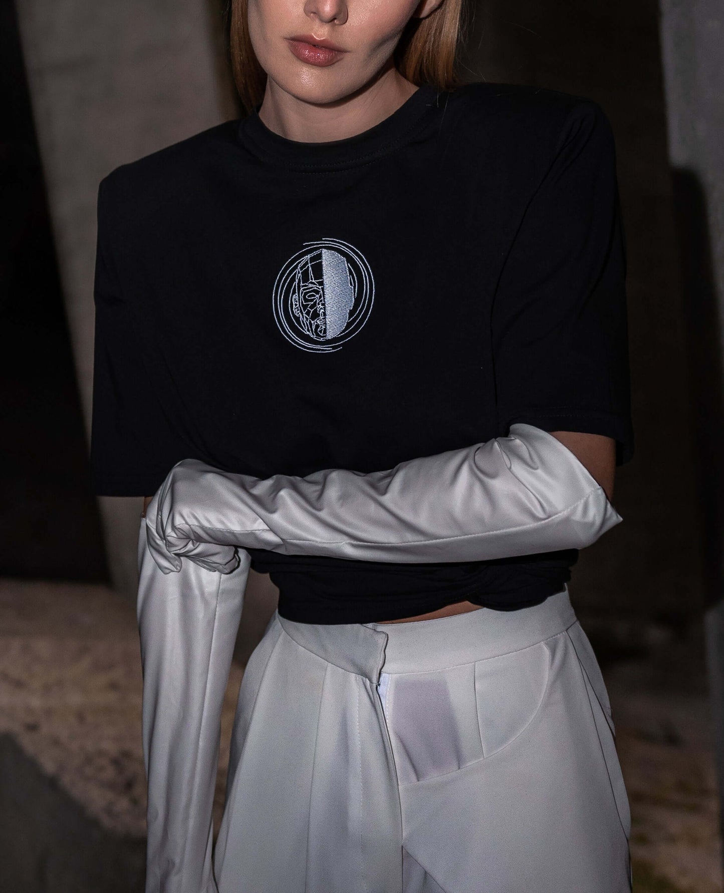 The model wears a black, oversized silhouette t-shirt with face embroidery in white. The outfit also contains long trousers and gloves, complimenting the black t-shirt with white spiral embroidery. The footwear is not visible in this photograph. An editorial photograph in an architectural monument in Plovdiv, Bulgaria.