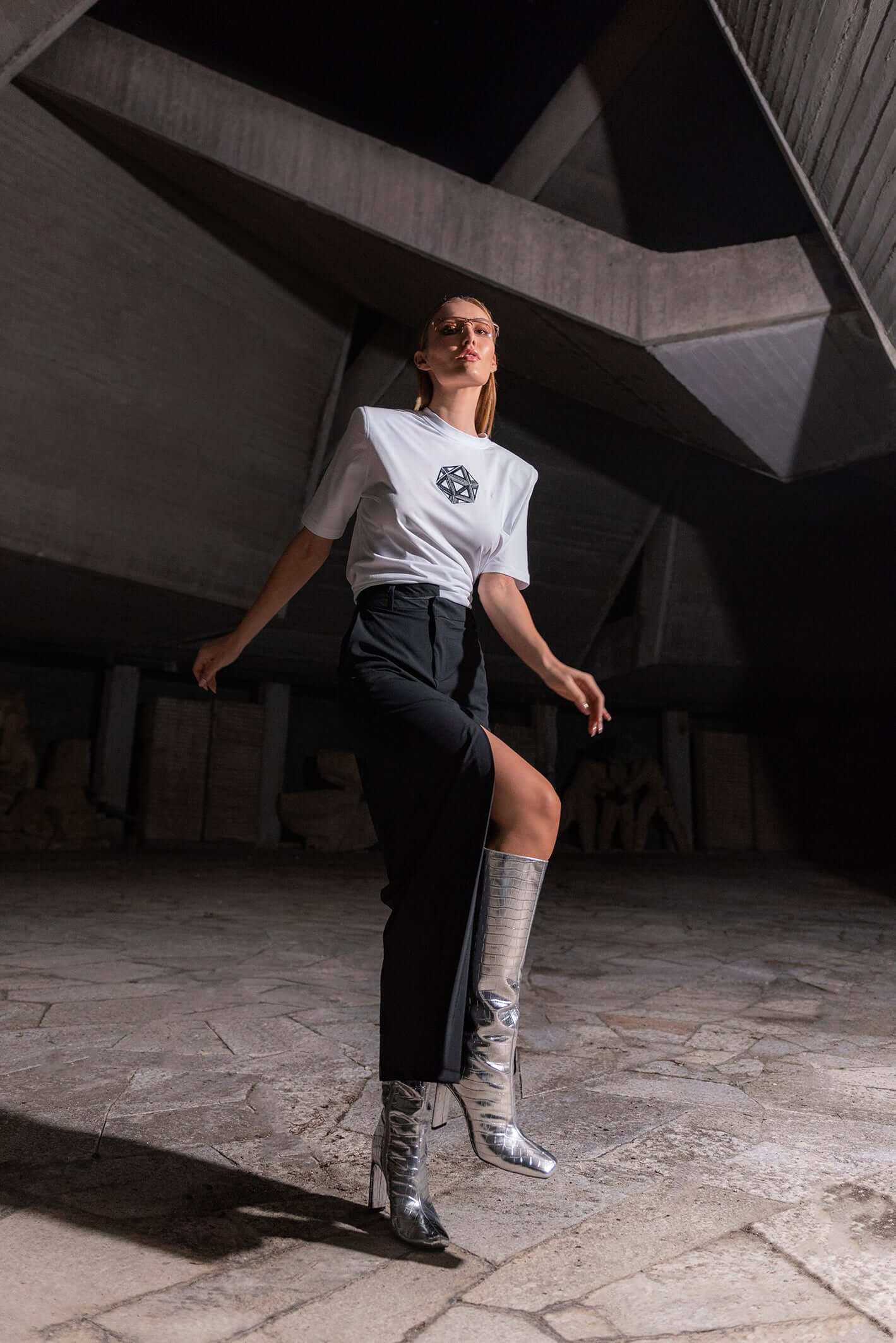 The model wears a white, oversized silhouette t-shirt with ICOSahedron embroidery in black. The outfit also contains a long skirt with a slit, boots and sunglasses, complimenting the white t-shirt with ICOSahedron embroidery. An editorial photograph in an architectural monument in Plovdiv, Bulgaria.