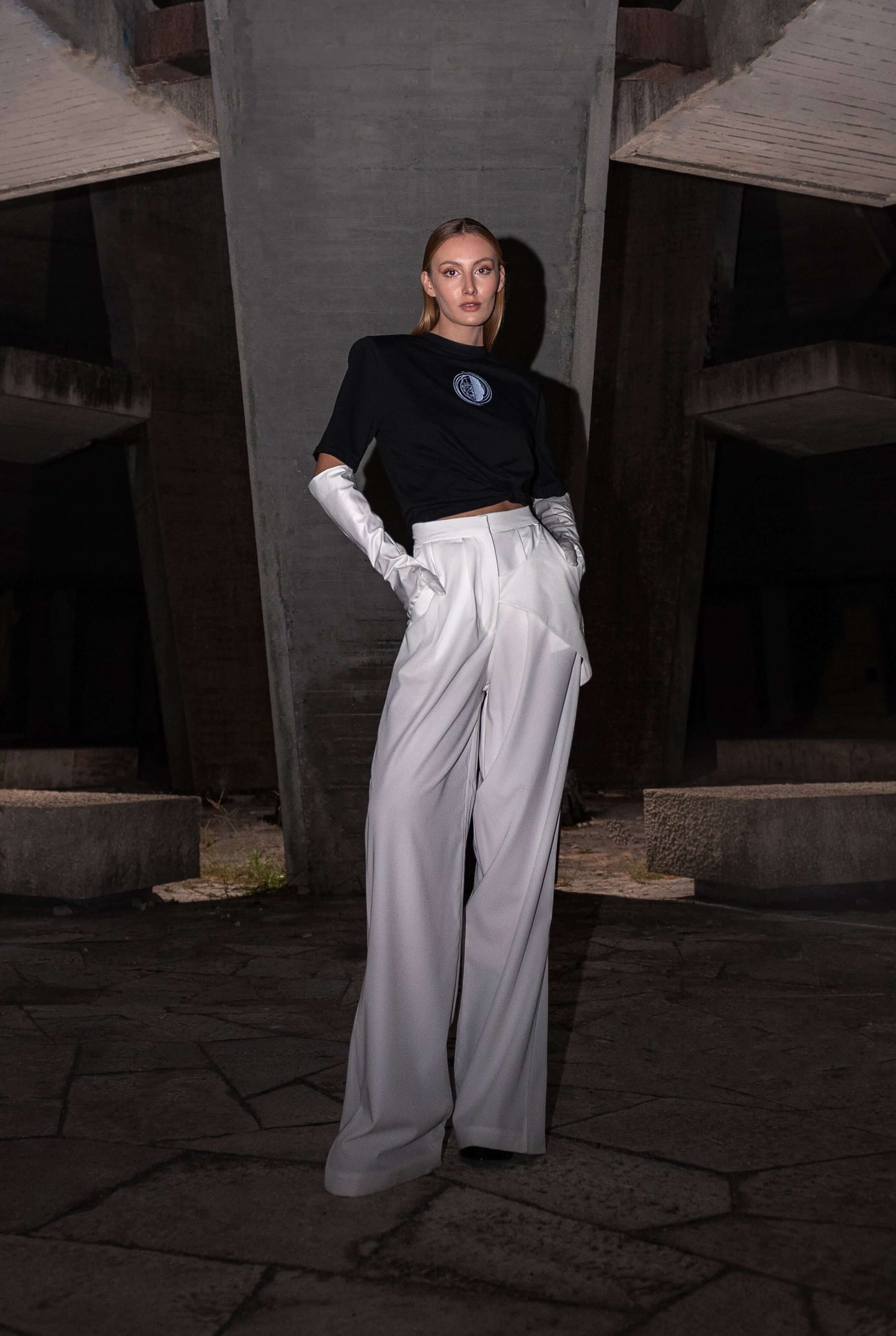 The model wears a black, oversized silhouette t-shirt with face embroidery in white. The outfit also contains long trousers and gloves, complimenting the black t-shirt with white spiral embroidery. The footwear is not visible in this photograph. An editorial photograph in an architectural monument in Plovdiv, Bulgaria.