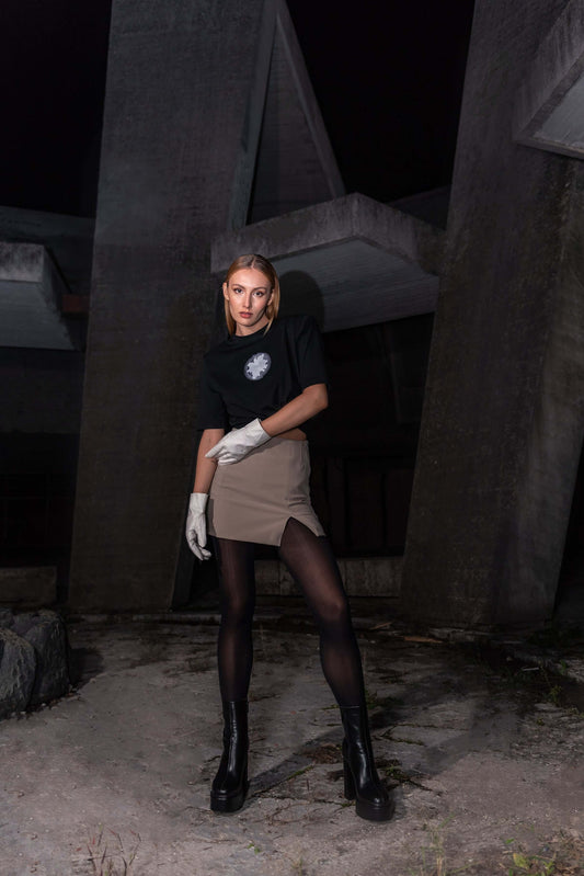 The model wears a black, oversized silhouette t-shirt with spiral embroidery in white. The outfit also contains a short skirt, gloves, tights and short, high-heel boots, complimenting the black t-shirt with white spiral embroidery. An editorial photograph in an architectural monument in Plovdiv, Bulgaria. 