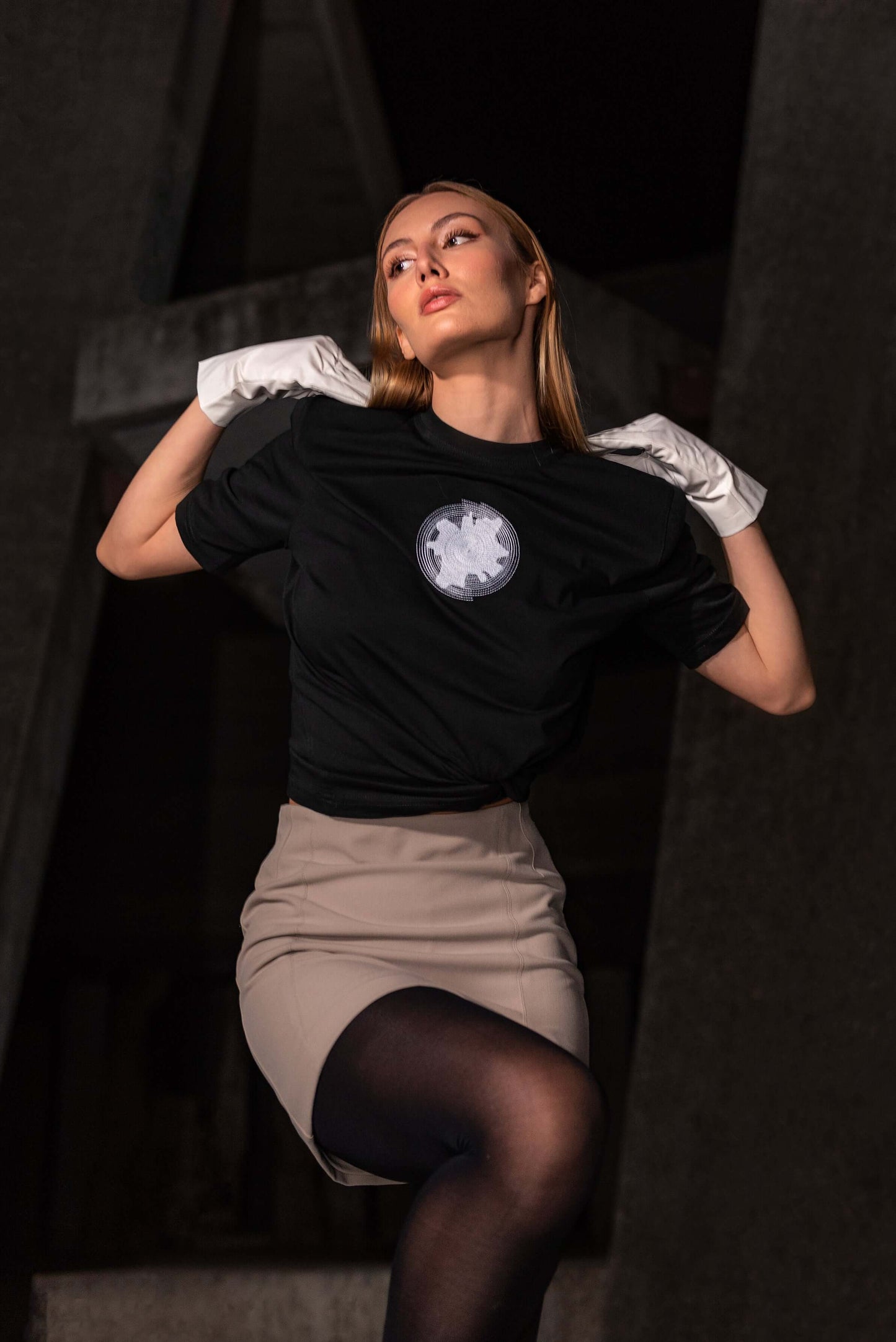 The model wears a black, oversized silhouette t-shirt with spiral embroidery in white. The outfit also contains a short skirt, gloves, tights and short, high-heel boots, complimenting the black t-shirt with white spiral embroidery. An editorial photograph of it in an architectural monument in Plovdiv, Bulgaria.