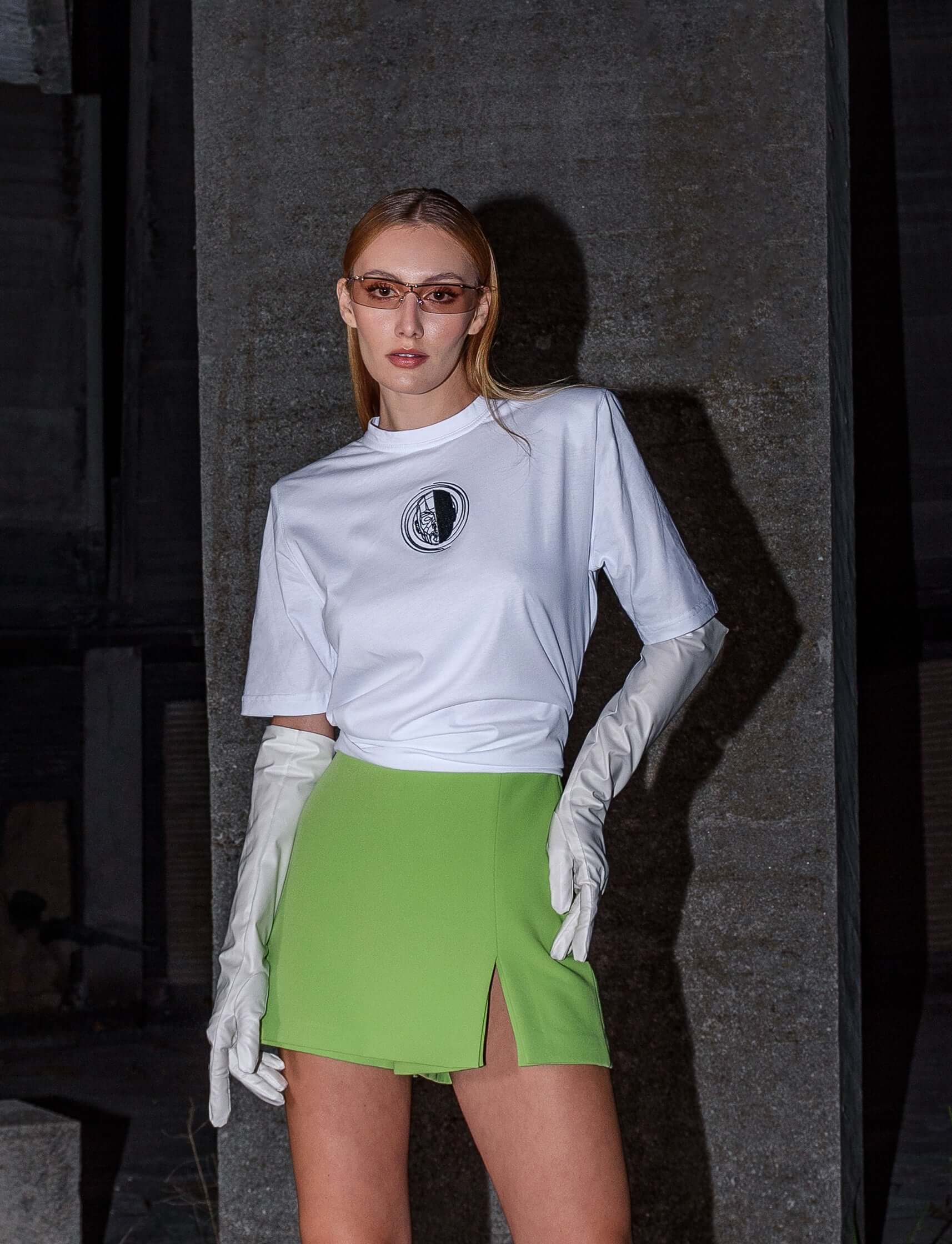 The model wears a white, oversized silhouette t-shirt with face embroidery in black. The outfit also contains a short skirt, boots and sunglasses, complimenting the white t-shirt with face embroidery. The footwear is not visible in this photograph. An editorial photograph in an architectural monument in Plovdiv, Bulgaria.