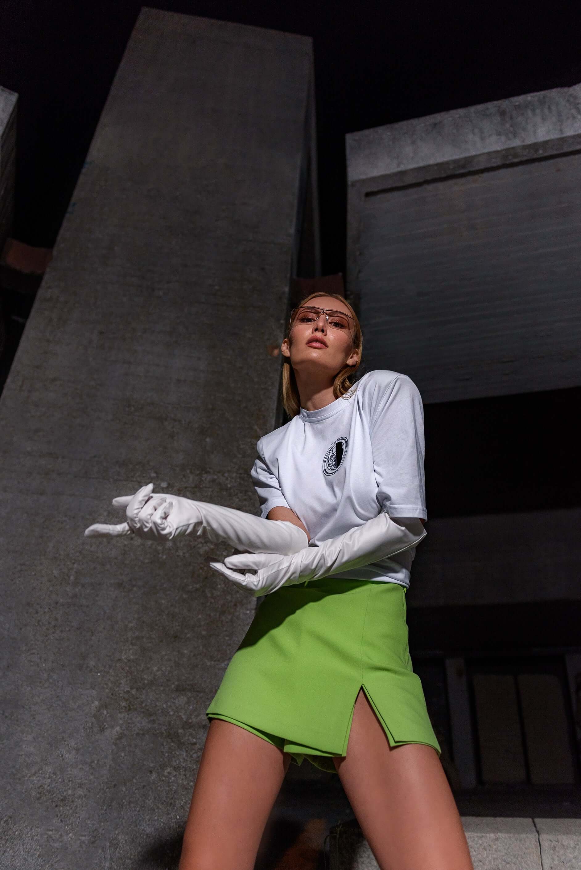 The model wears a white, oversized silhouette t-shirt with face embroidery in black. The outfit also contains a short skirt, boots and sunglasses, complimenting the white t-shirt with face embroidery. The footwear is not visible in this photograph. An editorial photograph in an architectural monument in Plovdiv, Bulgaria.