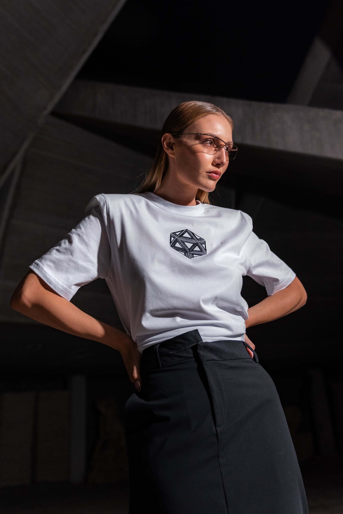 The model wears a white, oversized silhouette t-shirt with ICOSahedron embroidery in black. The outfit also contains a long skirt with a slit, boots and sunglasses, complimenting the white t-shirt with ICOSahedron embroidery. An editorial photograph in an architectural monument in Plovdiv, Bulgaria.