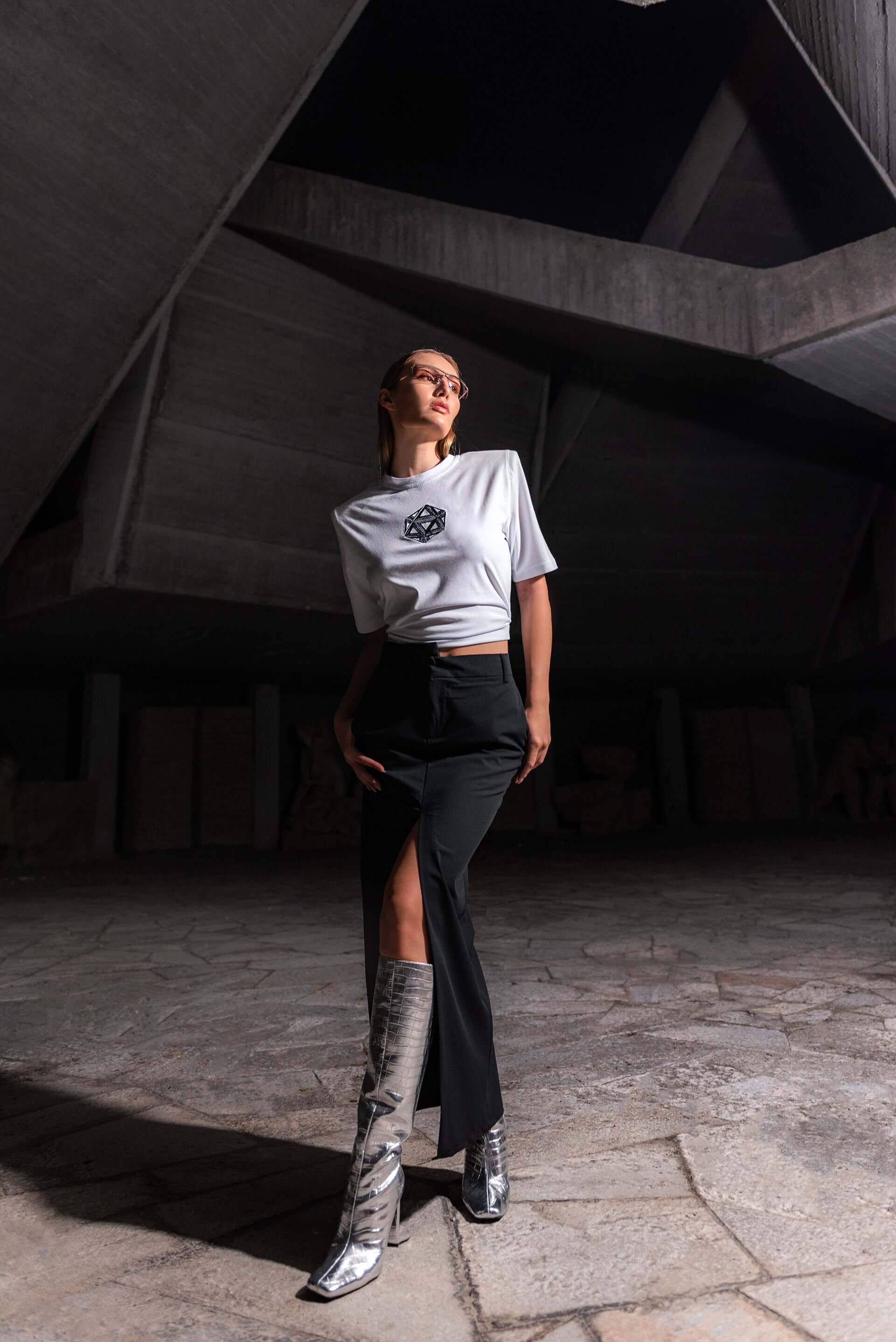 The model wears a white, oversized silhouette t-shirt with ICOSahedron embroidery in black. The outfit also contains a long skirt with a slit, boots and sunglasses, complimenting the white t-shirt with ICOSahedron embroidery. An editorial photograph in an architectural monument in Plovdiv, Bulgaria.